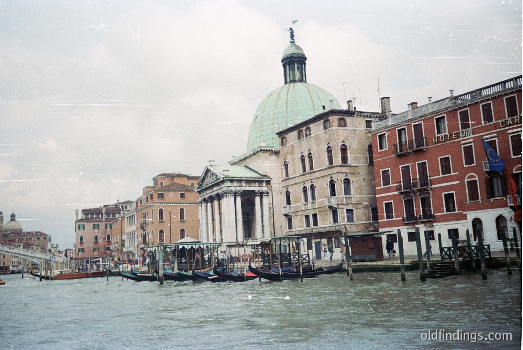 Venetian canal scene featuring the dome of **Santa Maria della Salute** and adjacent historic buildings. Traditional gondolas docked along wooden piers, with classic Venetian architecture in muted tones. Likely late 20th century,