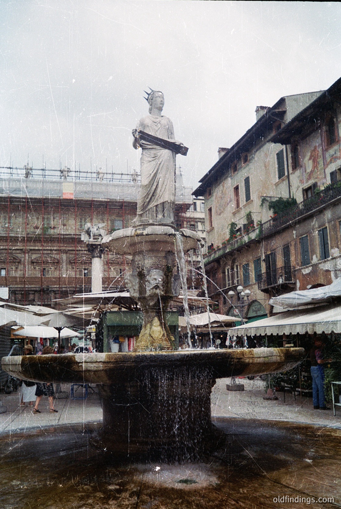 Classic Renaissance-style fountain featuring a robed female figure atop a multi-tiered basin in an Italian piazza. Surrounding buildings exhibit arched windows and weathered stucco. Market stalls with umbrellas line the square, hinting at a lively public space. Likely Verona, Italy (, , , , )