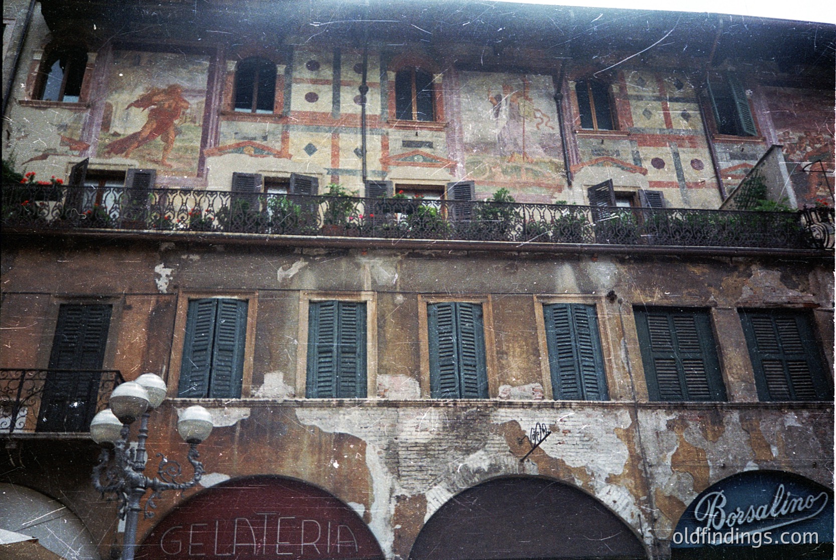 Faded Renaissance-style building façade featuring frescoed upper balcony with mythological figures, aged plaster, and arched ground-level entrances. Signage includes "Gelateria" and "Borsalino." Likely Italy, 19th–early 20th century.
