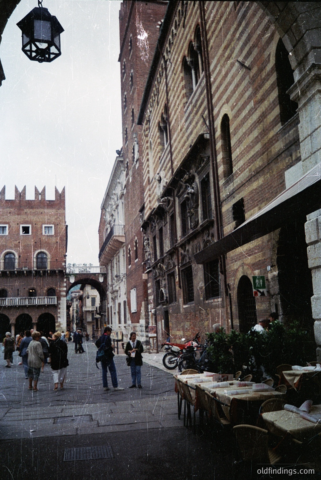 Venetian alleyway showcasing historic brick facades with arched windows and balconies. Cobblestone street lined with outdoor café tables and vintage lanterns. Crowd of casually dressed pedestrians in a narrow urban setting.