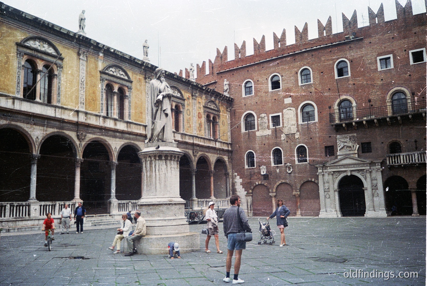 Historic Piazza del Capitolo in **Siena, Italy**, featuring Gothic architecture with crenellated roofline and arched colonnades. Central equestrian statue on a pedestal dominates the square. Crowd of casually dressed people, including a cyclist and seated individuals, suggests mid-20th century (likely 1960s-1970s) daily life.