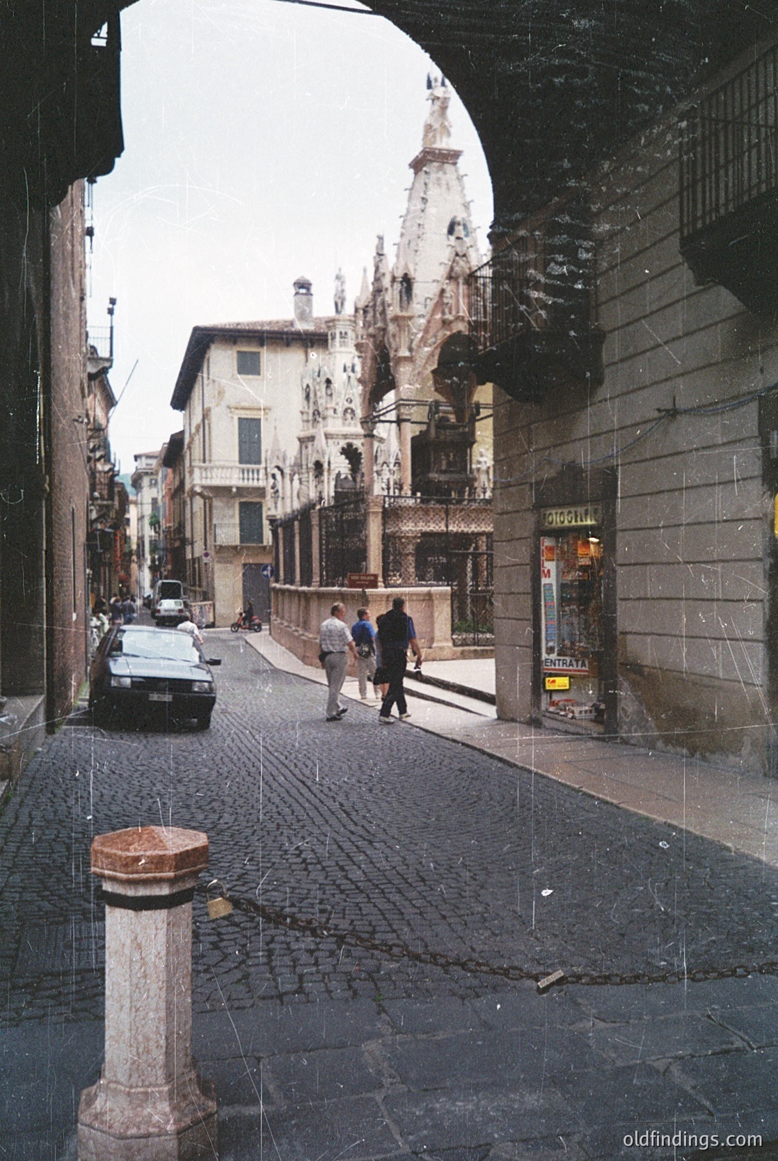 Historic European street scene under a stone archway, likely (). Cobblestone pavement, vintage car, and Gothic-style buildings with ornate facades frame the view. Two pedestrians walk toward a grand cathedral spire in the distance. Raindrops add texture to the scene, evoking mid-20th century ambiance.