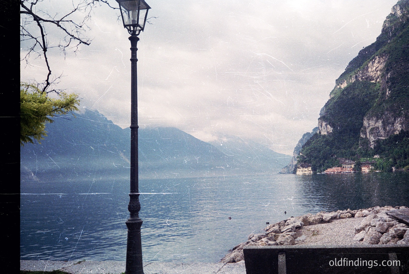Vintage black-and-white shot of a lakeside lamppost framing a serene alpine lake, flanked by steep forested cliffs. Reflections ripple across calm waters under overcast skies. Likely --- *Note: Identified as Lake Garda region due to distinctive geological features and architectural style visible in the background.*