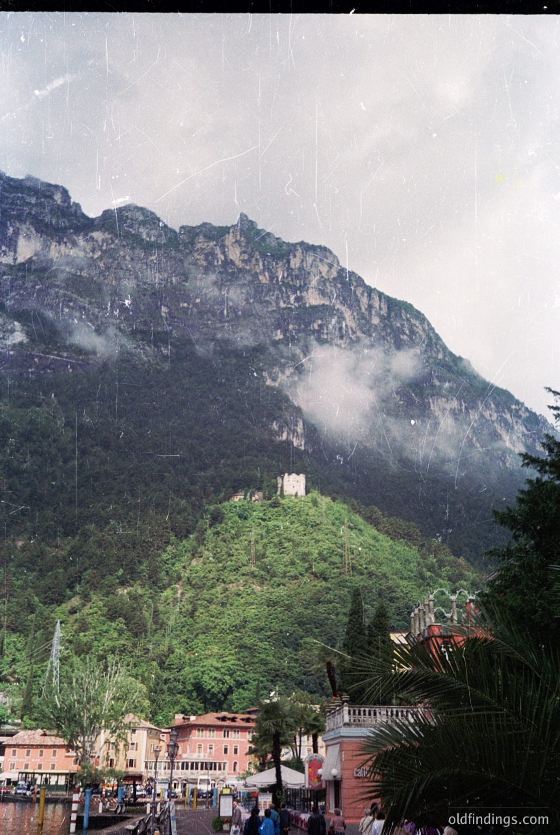 Historic castle perched atop lush green hillside, framed by dramatic alpine peaks. Foreground features Mediterranean-style buildings with palm trees, suggesting a European alpine resort. Overcast skies enhance the rugged mountain backdrop. Likely European architecture in a mountainous region.