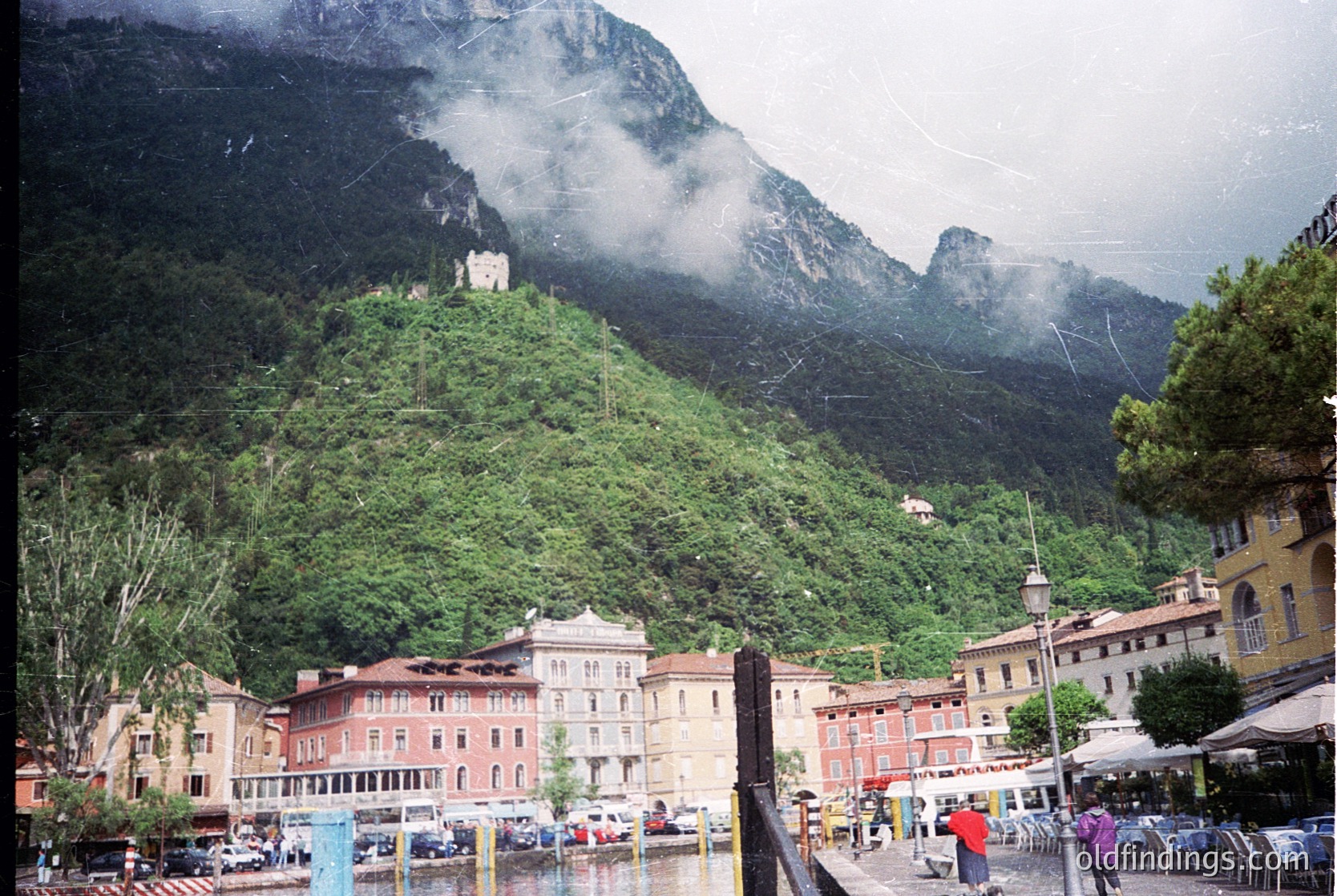 Alpine lakeside village nestled between dramatic rock formations and lush greenery. Colorful, multi-story buildings with European architectural details line the waterfront promenade, featuring balconies and pastel facades. A historic stone castle perched atop a hill dominates the background. Outdoor seating and pedestrians suggest a bustling yet serene tourist hub.