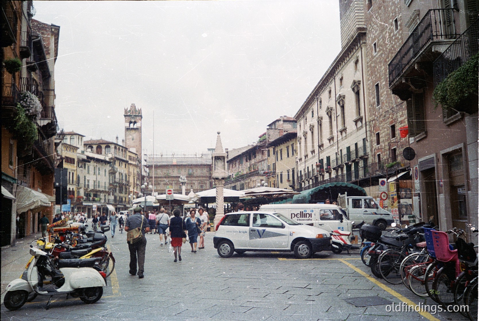 Vibrant 1990s-era street scene in **Bologna, Italy**, showcasing classic medieval architecture with arched doorways and brick facades. Pedestrians, scooters, and a white taxi with "Pellini" branding navigate the cobblestone street lined with outdoor market stalls. The distant **Bologna Town Hall** (Piazza Maggiore) and **Two Towers** frame the scene.