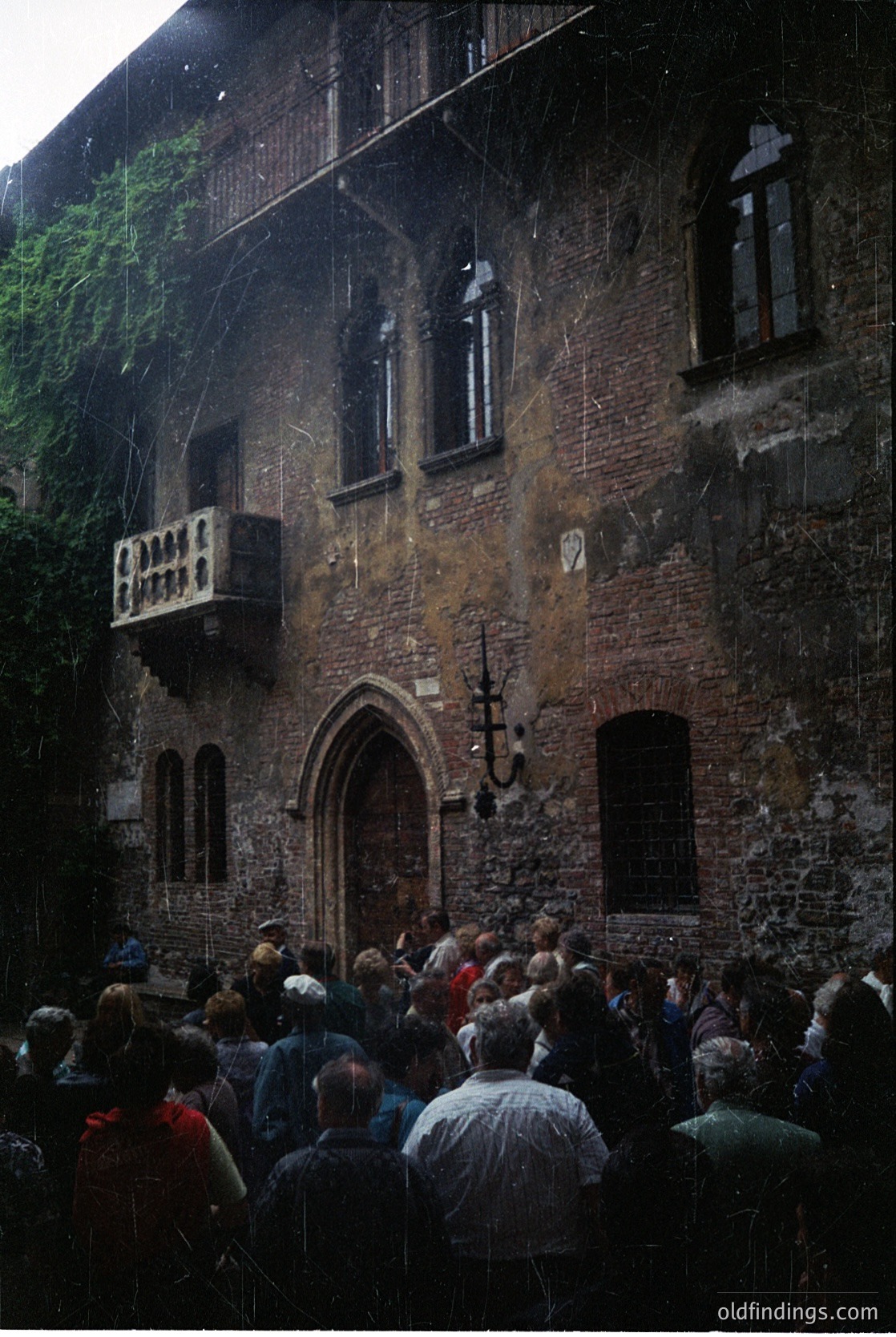 Historic stone building with Gothic Revival arches, balconies, and wrought-iron lanterns. Crowd gathers at arched entrance, suggesting cultural or tourist significance. Weathered brickwork and ivy-clad walls indicate age. Likely European, possibly or early .