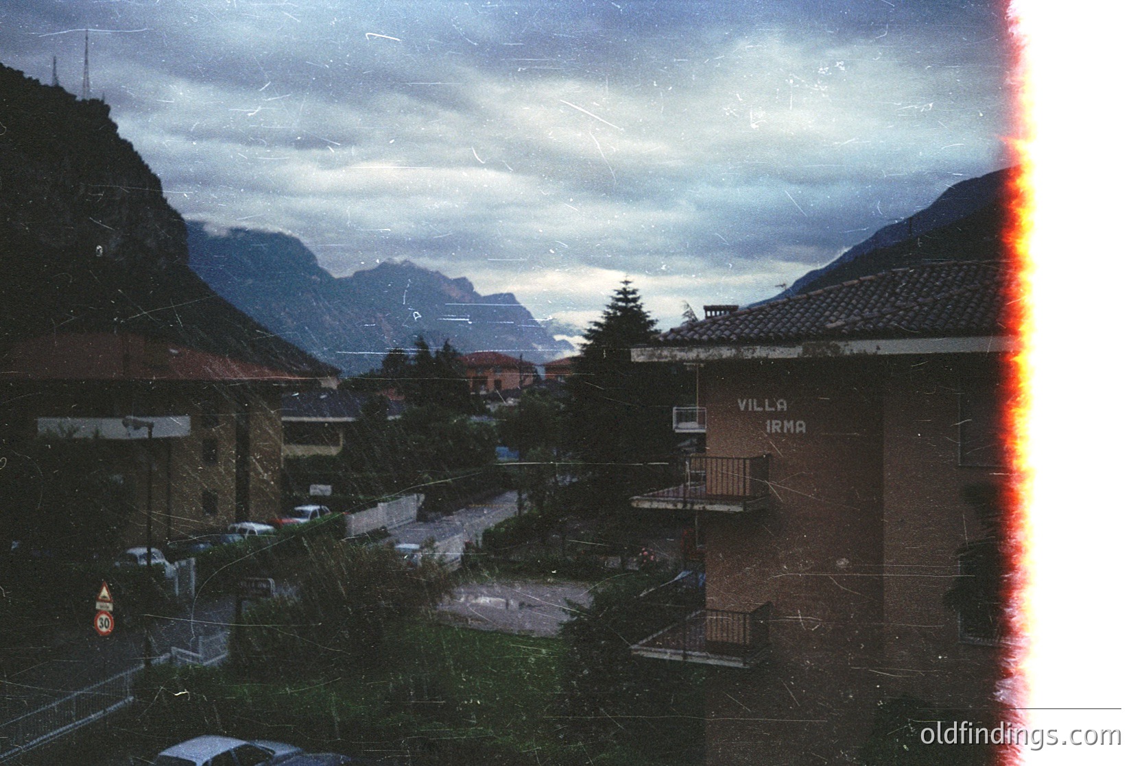 Vintage alpine village scene with "Villa Irma" signage, framed by aged photo borders. Snow-capped peaks loom under stormy skies, while brick buildings and parked cars suggest mid-20th-century European mountain resort.