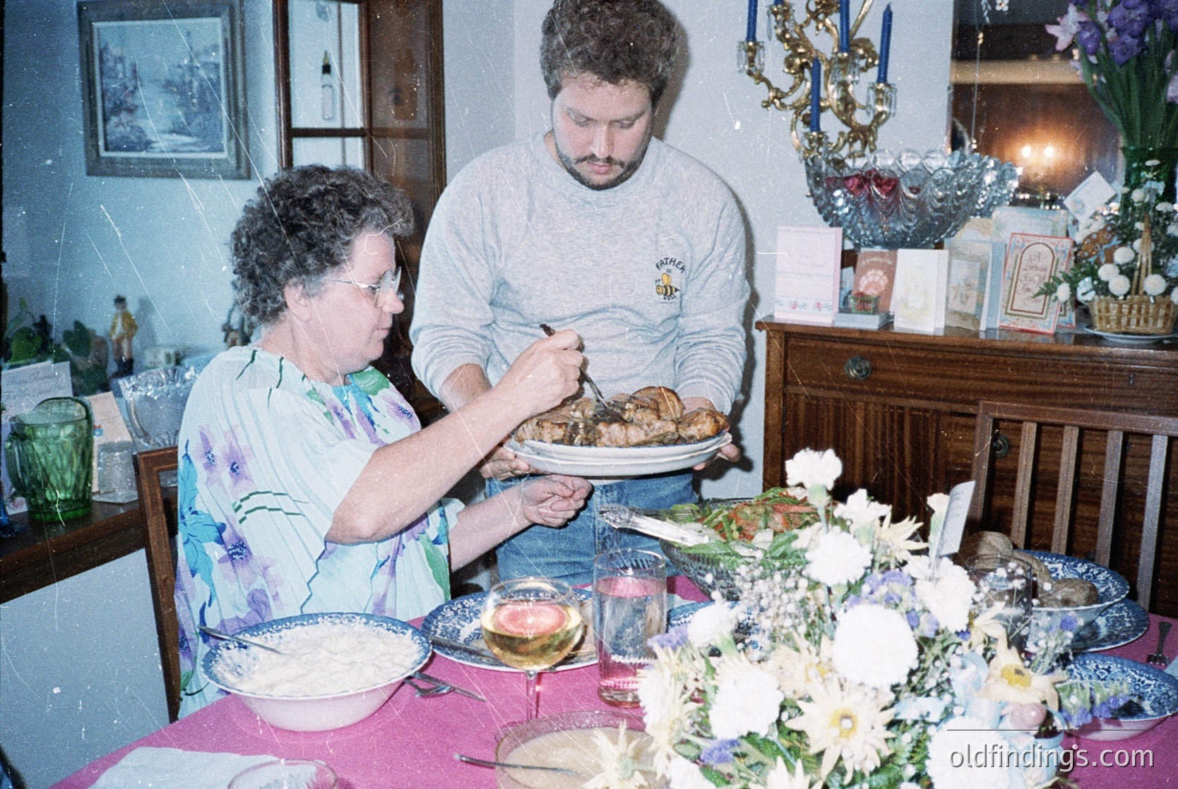 Vintage indoor family meal scene: woman and man serving roasted chicken at a floral-decorated table with vintage dishes. Wooden cabinet, potted plants, and holiday garland suggest late 20th-century domestic life.