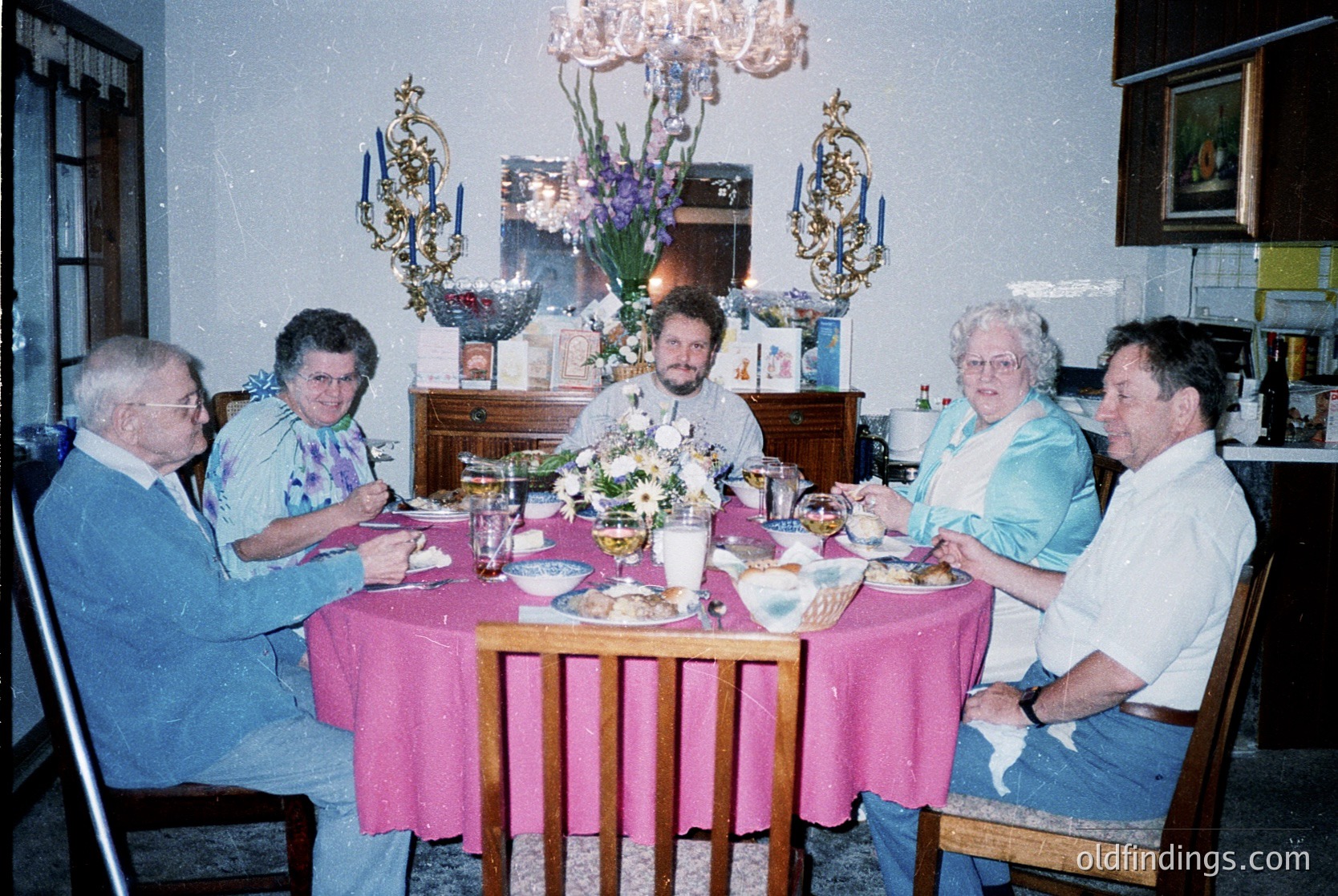 Family gathering at a mid-century dining table, adorned with floral centerpieces and vintage chandelier. Six adults seated in wooden chairs, dressed in 1980s-90s casual attire (blouses, button-downs, jeans). Indoor setting with floral wallpaper and framed art. Warm, nostalgic domestic scene.