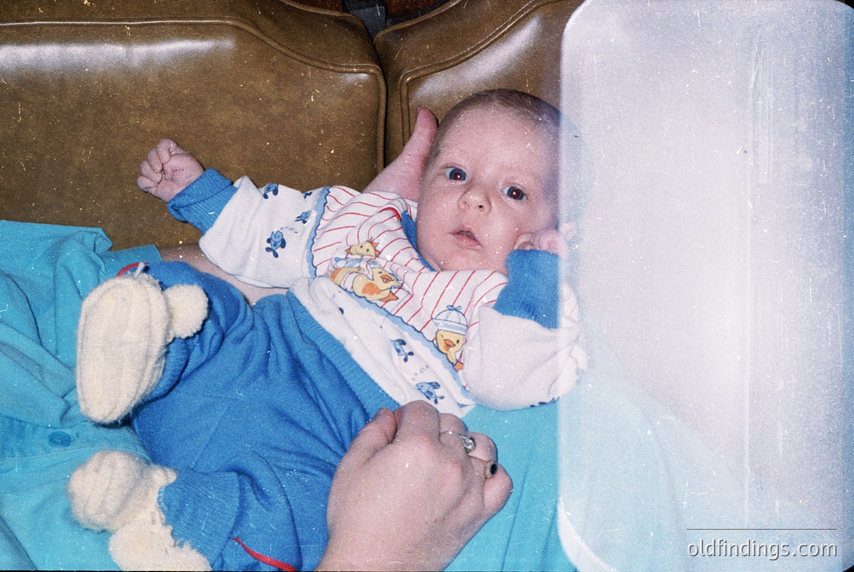 Vintage-style photo of an infant in a hospital bassinet, wearing a striped onesie and holding a white teddy bear. Soft focus and warm sepia tones suggest 1980s–1990s medical photography.