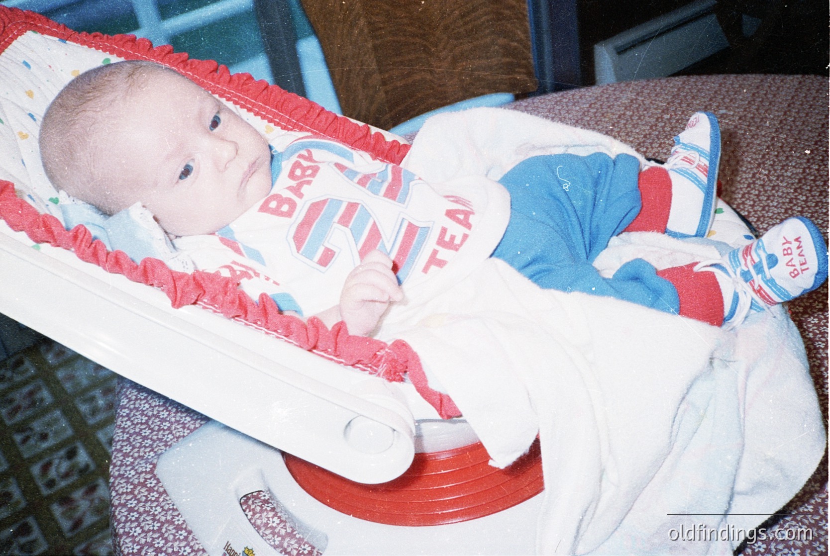 Vintage baby in a red-and-white "Baby Team" highchair, wearing matching onesie and socks. Indoor setting with patterned upholstery. Likely late 20th century (1980s-1990s).