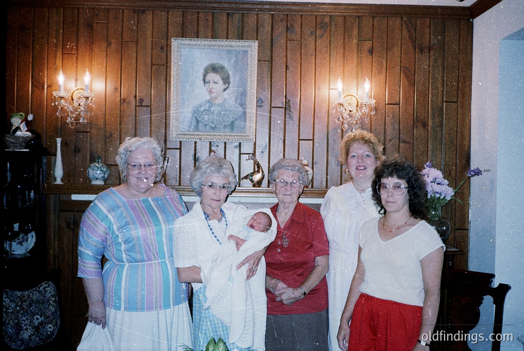 Family portrait featuring five women in a rustic interior, likely 1980s–1990s. Central figure holds a newborn, surrounded by four smiling elders in casual attire (striped dress, red sweater, white blouse, red skirt). Wood-paneled walls, vintage candelabras, and a framed portrait add warmth. Possible celebration or milestone occasion.