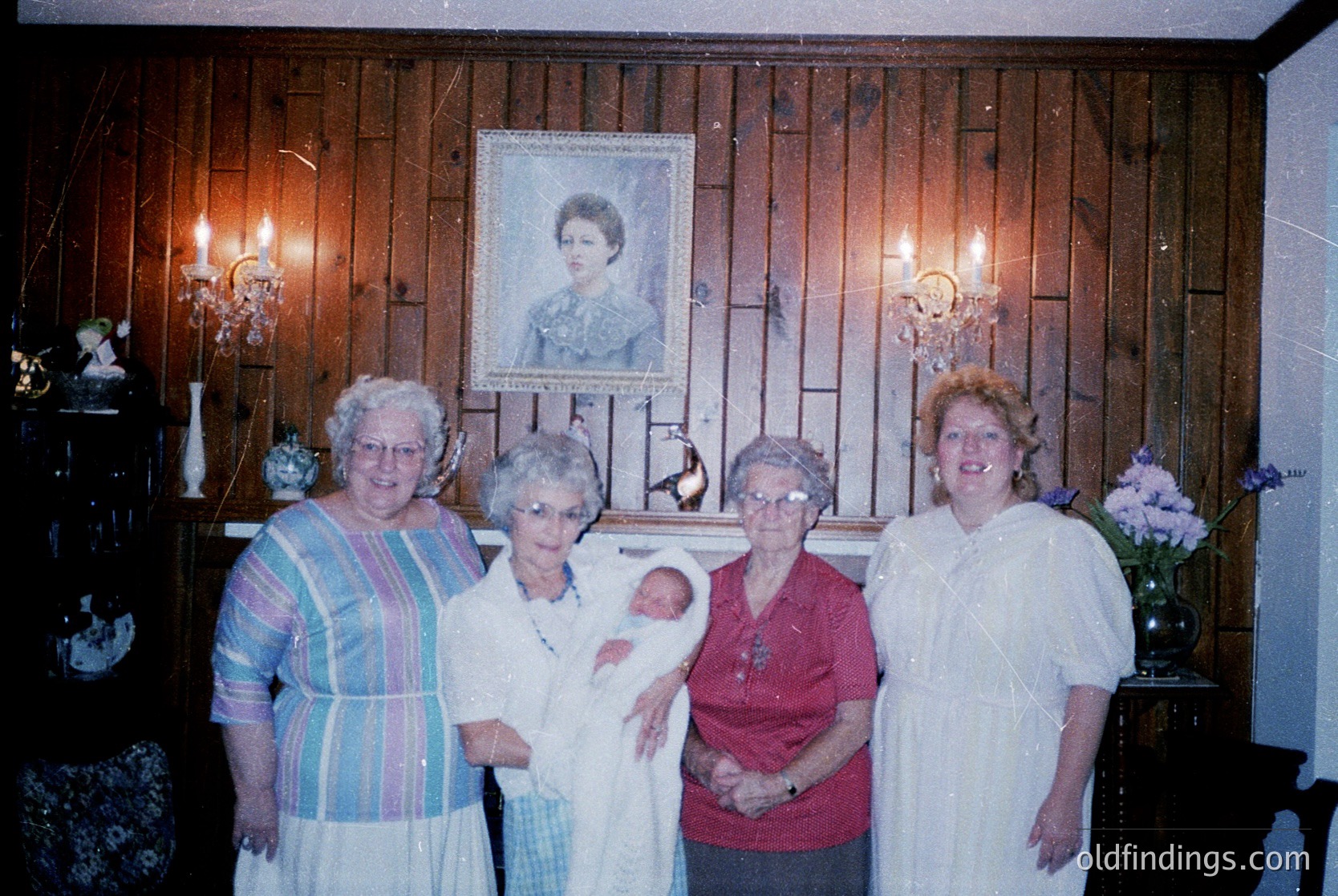 Four women pose indoors in a vintage-style setting, likely 1980s–1990s. Wood-paneled walls feature framed portraits, candle sconces, and floral arrangements. The central woman holds a baby, while others wear striped, floral, and solid blouses. Warm lighting enhances the nostalgic ambiance.