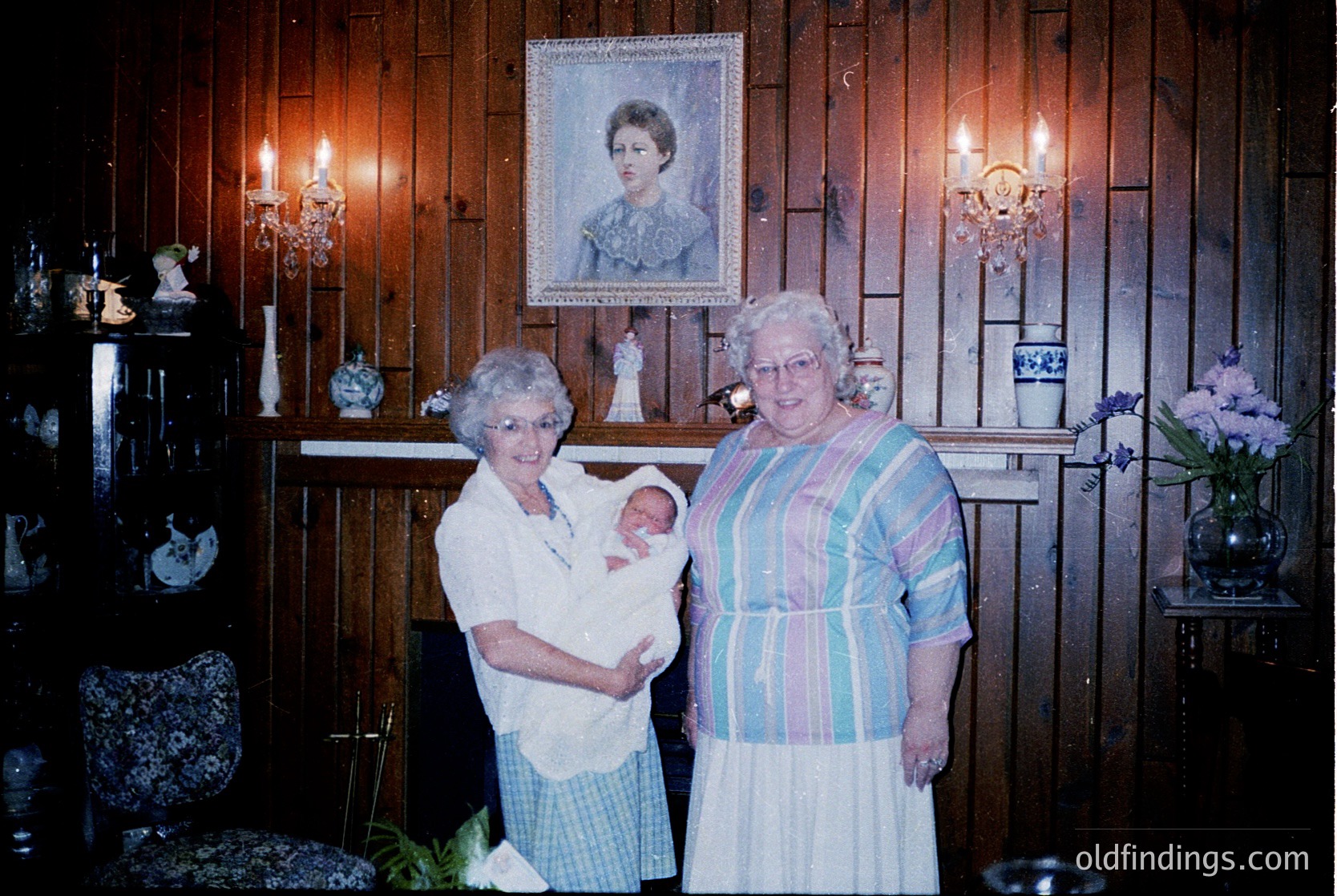 Two elderly women pose indoors with a newborn in a vintage-style home, likely 1970s–1980s. Wood-paneled walls, ornate wall sconces, and a framed portrait above a mantelpiece with decorative figurines and floral arrangements. One woman wears a striped dress; the other, a white blouse with a floral skirt.