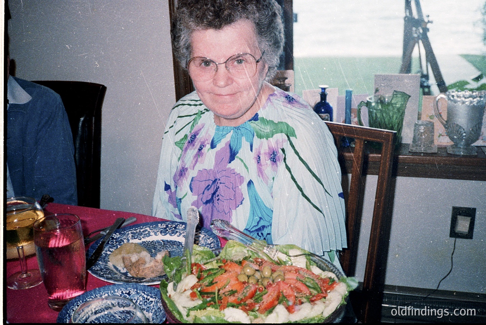 Vintage indoor portrait of a woman in a floral blouse, seated at a table with a large salad and blue-and-white dishware. Glasses, a pitcher, and a wall socket suggest mid-20th century domestic setting.