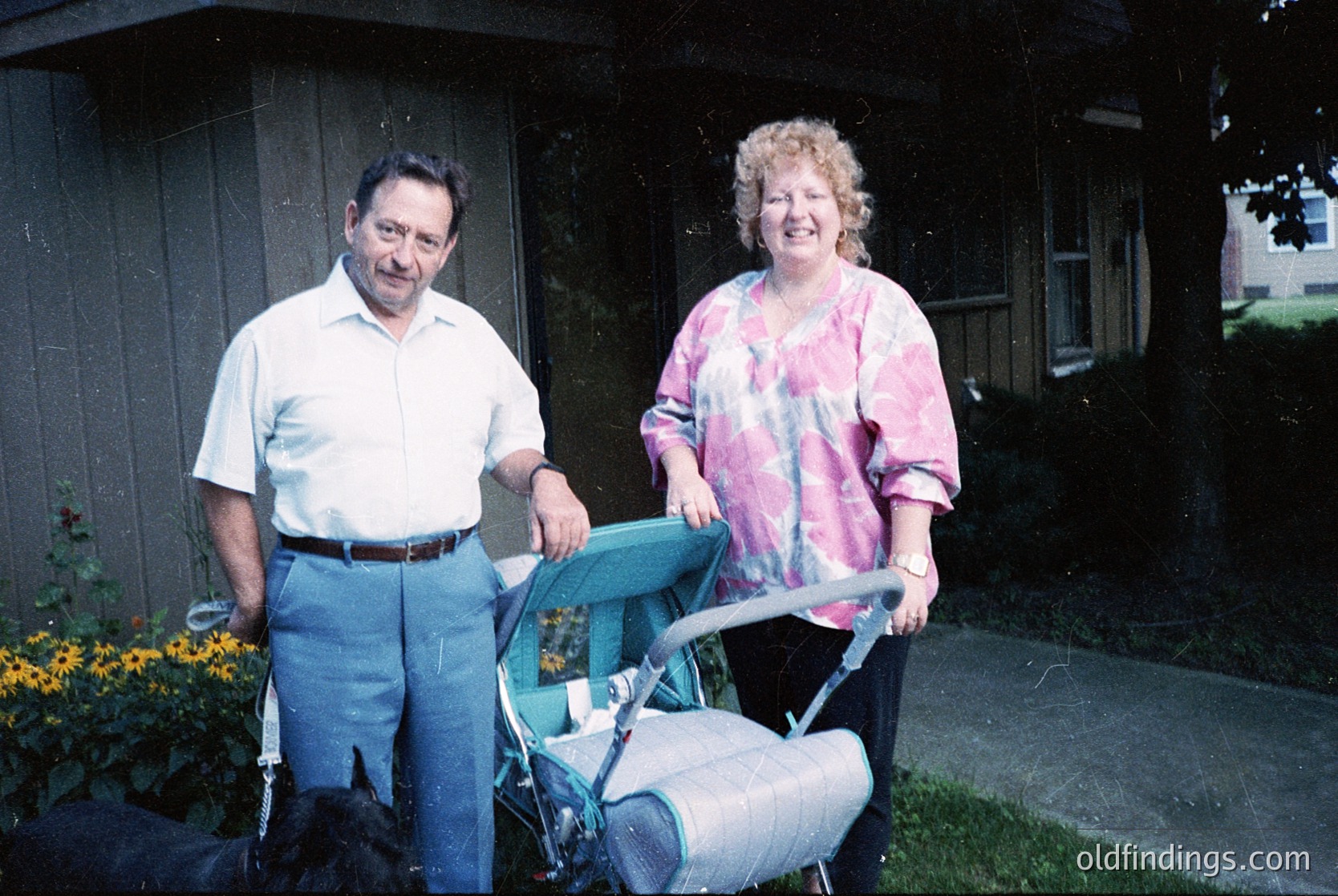 Mid-20th century suburban portrait: man in light button-down shirt and dark trousers, woman in tie-dye blouse, posing with a vintage stroller on a paved driveway. Residential home with brick accents and flowering shrubs in background. Likely 1970s–1980s USA.