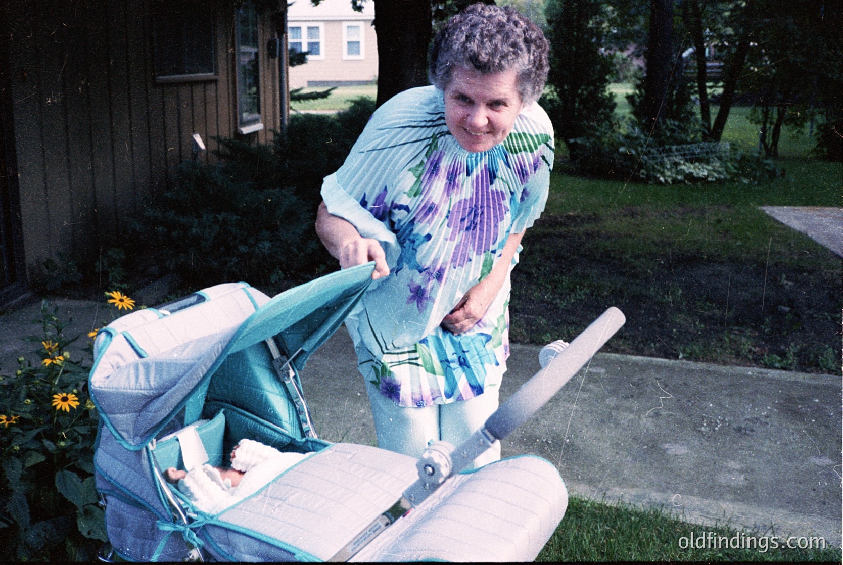Vintage 1970s suburban scene: woman in patterned blouse and floral skirt adjusts a classic green-and-white stroller with a baby inside. Residential backyard with yellow flowers and brick pathway.