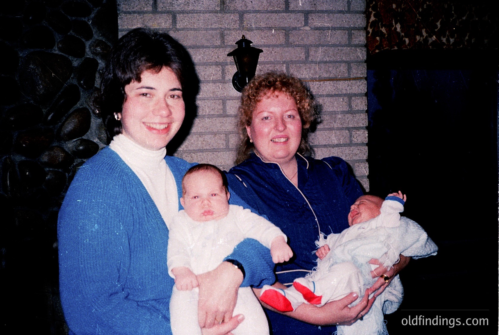 Two women pose indoors with infants, 1980s-1990s style. Left: woman in blue sweater, dark glasses, holding baby in white onesie. Right: woman in blue top, curly hair, cradling baby in white bib and red diaper. Brick wall, vintage lantern, and floral wallpaper visible.