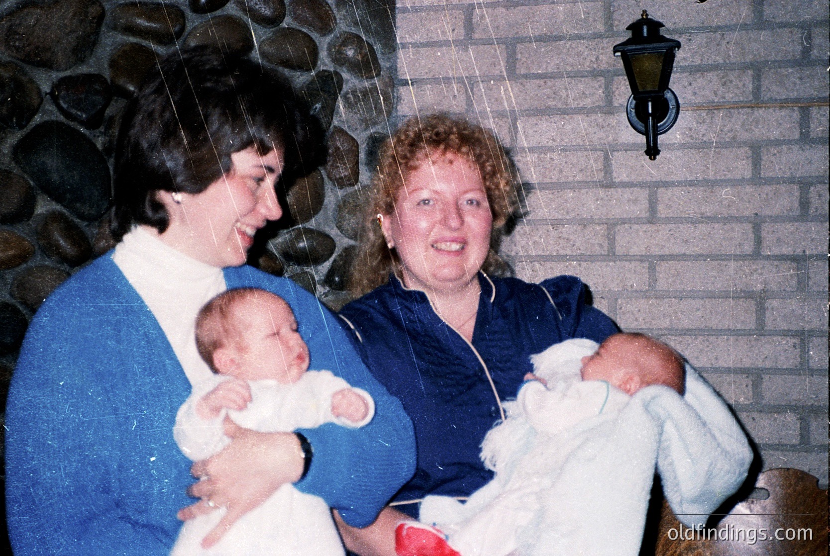 Two women holding infants in a cozy indoor setting with stone wall and vintage lamp. Likely 1980s–1990s family portrait. Warm, nostalgic atmosphere.