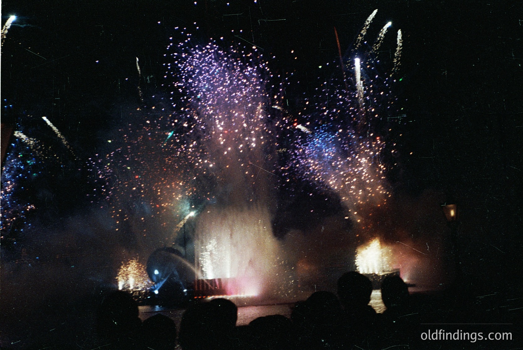 Fireworks display illuminating night sky with vibrant bursts of purple, blue, and gold. Silhouetted crowd faces forward, capturing communal celebration. Dynamic motion blur highlights explosive energy. Ideal for festive, patriotic, or seasonal stock imagery.