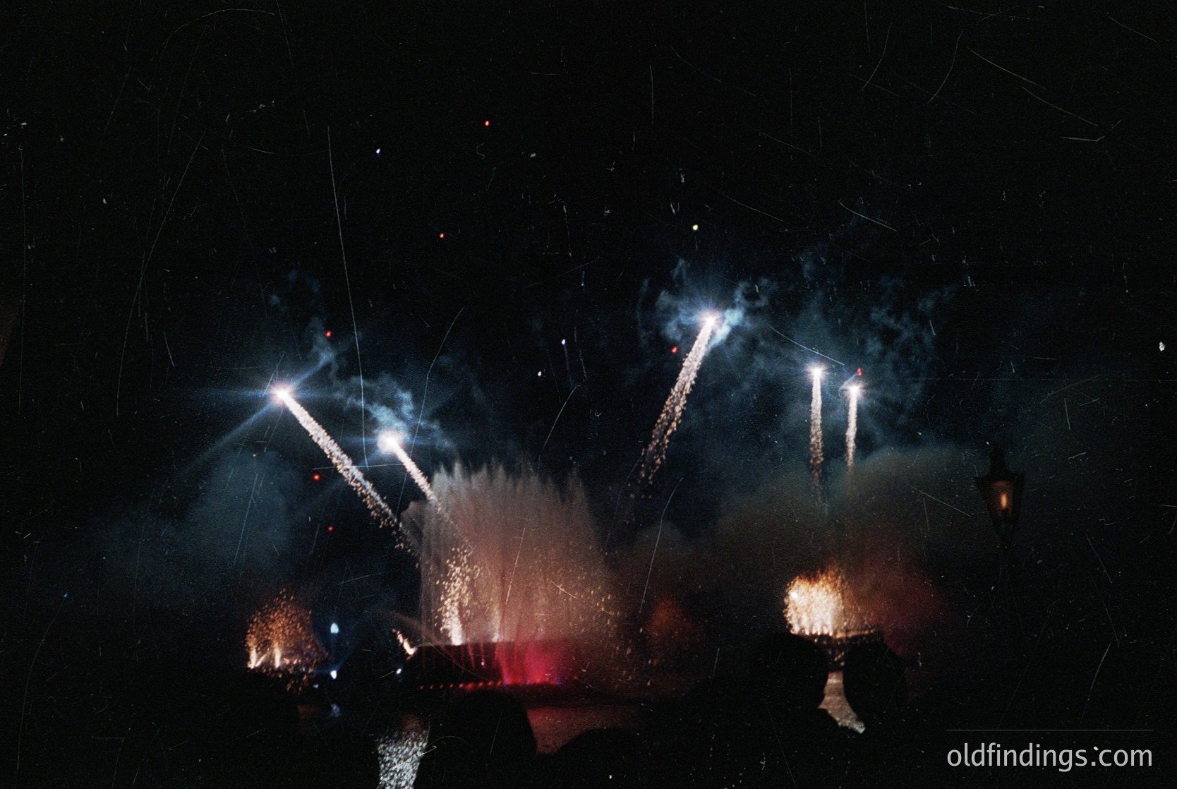 Fireworks display illuminating night sky with starbursts and cascading trails. Silhouetted figures below suggest crowd celebration. Dynamic motion blur captures explosive energy. Likely urban festival or holiday event.