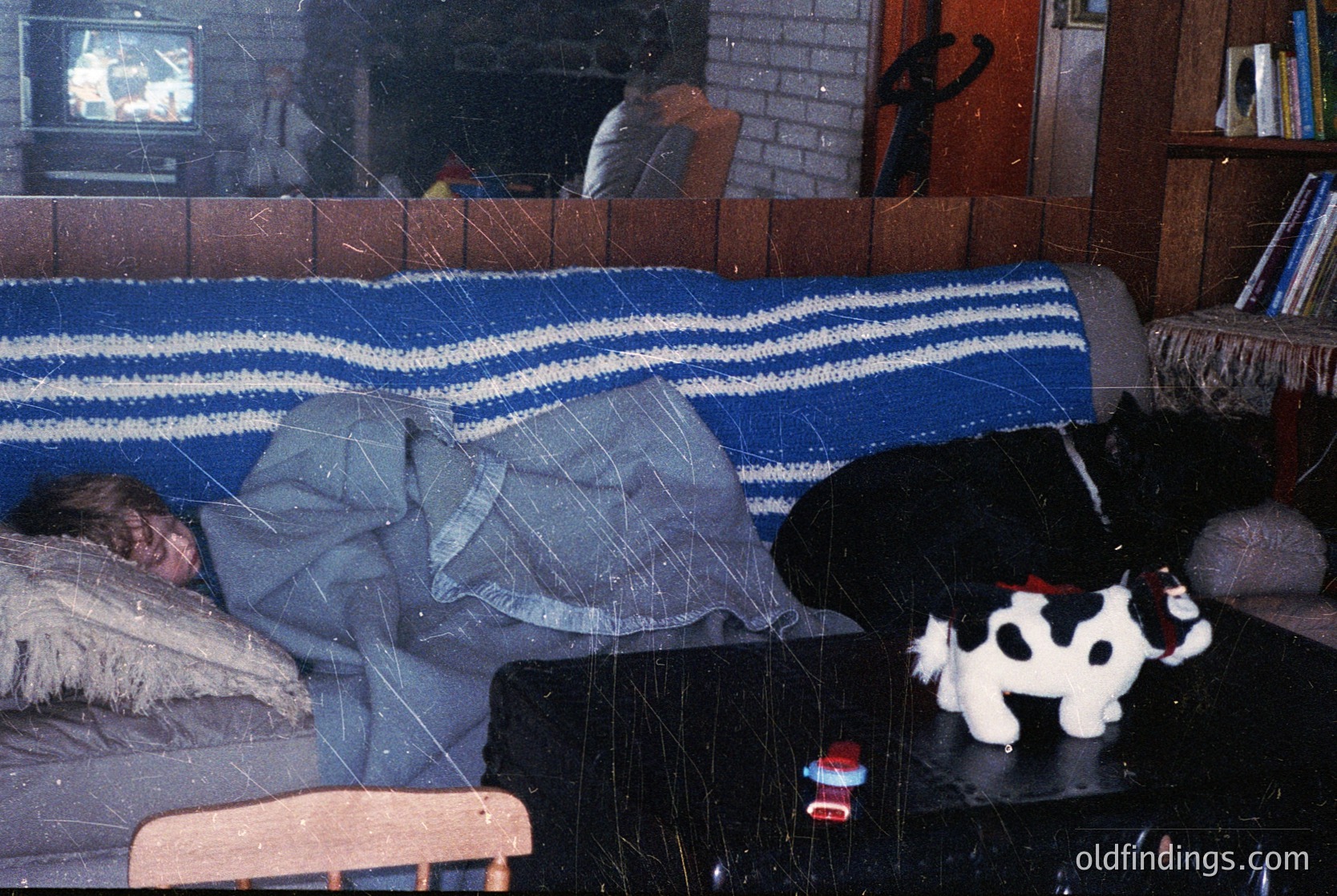 Vintage living room scene with a child sleeping on a striped blanket-covered sofa, surrounded by retro decor. A black-and-white cow plush toy sits nearby, while a CRT TV broadcasts static. Brick fireplace and wooden furniture suggest mid-20th-century domestic life.