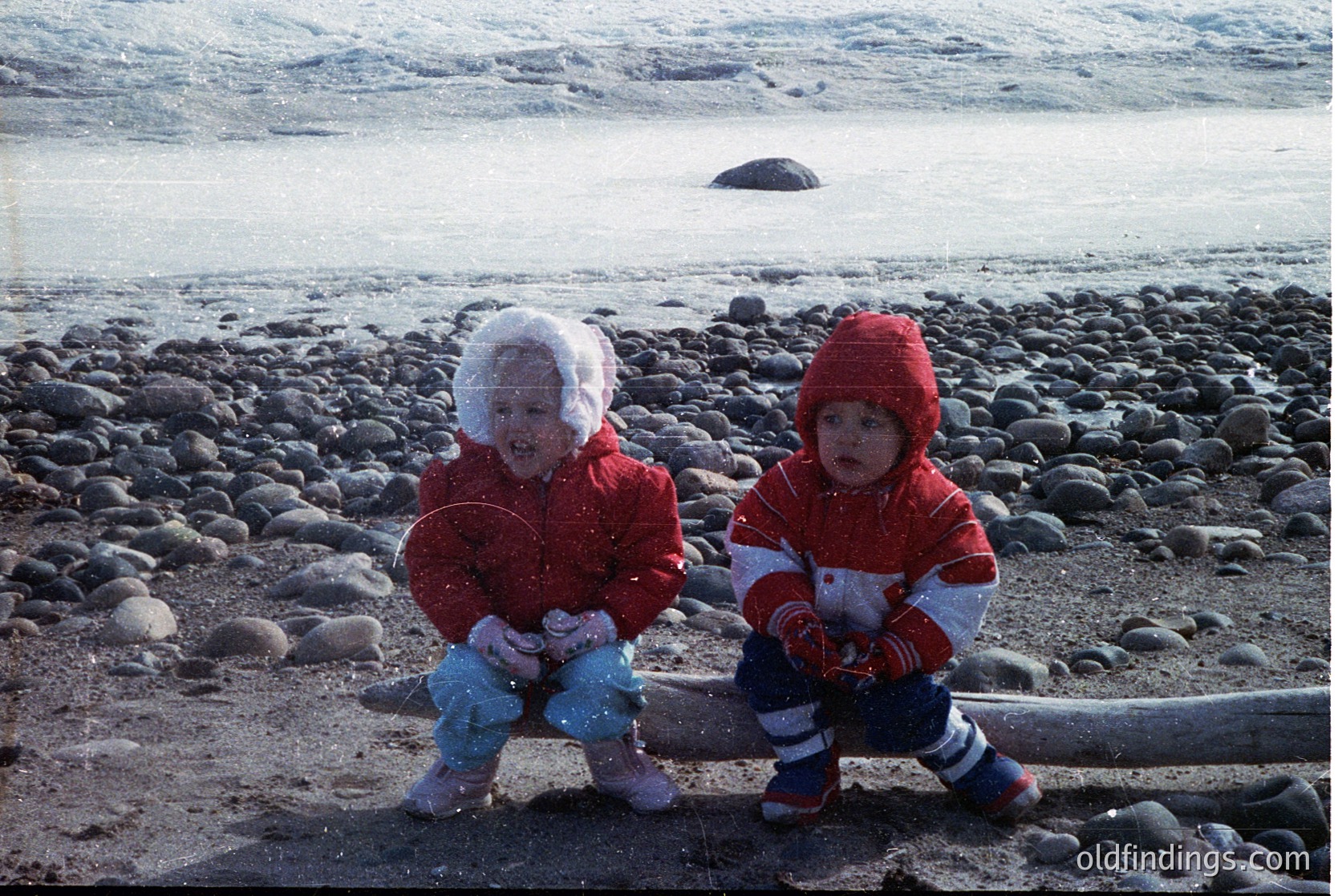Two children in winter attire sit on rocky shoreline, playing in light snowfall. Both wear red jackets, hats, and mittens; one has blue pants, the other striped. Coastal rocks and distant waves form a serene winter seascape. Likely 1980s–1990s, Northern Hemisphere.