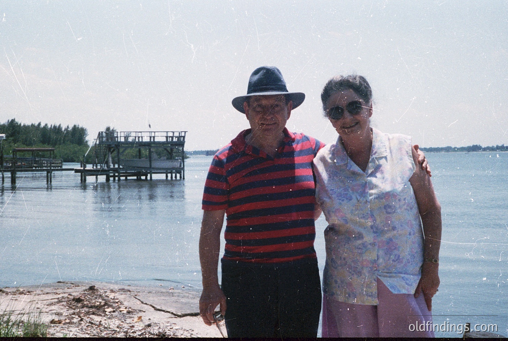 Couple posing by a lakeside dock in retro summer attire—man in striped shirt & fedora, woman in floral blouse & sunglasses. Mid-20th century (1960s-70s) beach or lakeside scene with wooden pier and greenery. Vintage photography with slight grain.