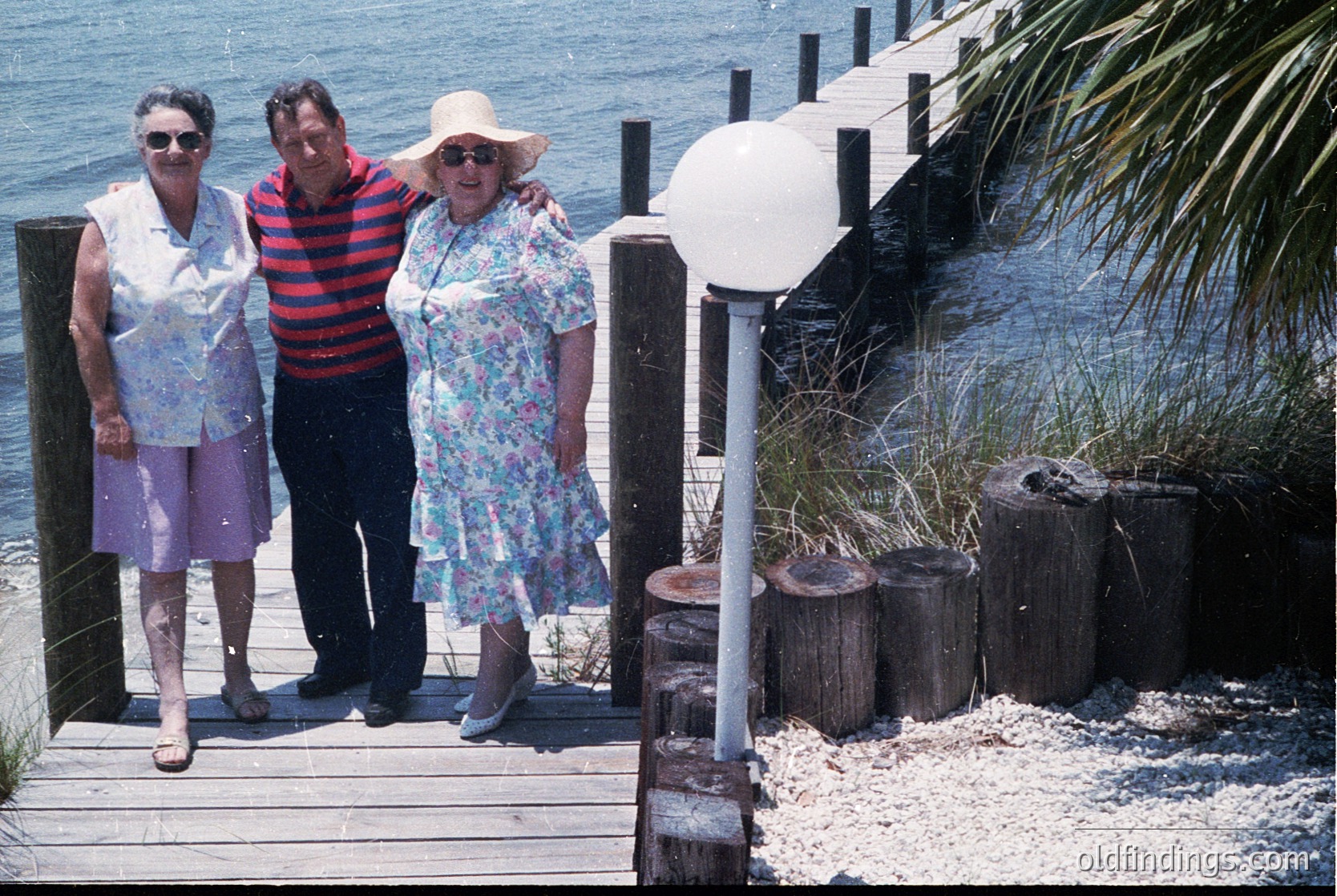 Three adults pose on a wooden pier by the sea, 1970s-1980s. Woman left wears patterned blouse, purple pants, and sandals; man in red/white striped shirt; woman right in floral dress, hat, and sandals. Pier features rustic wooden railings and lamppost. Coastal vegetation and pebbled shoreline visible.