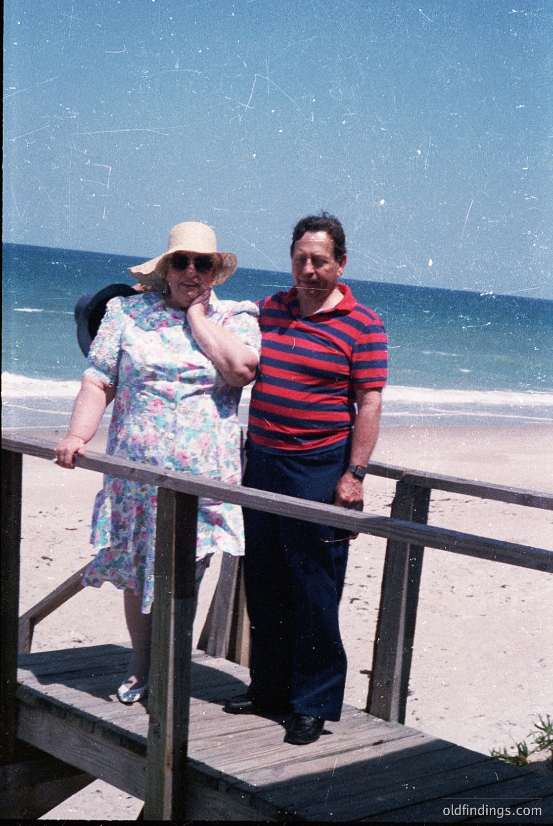 Vintage seaside couple posing on a wooden pier, 1960s-70s. Woman in floral dress, straw hat, and sunglasses; man in red-and-white striped polo. Clear blue ocean and sandy shore in background.