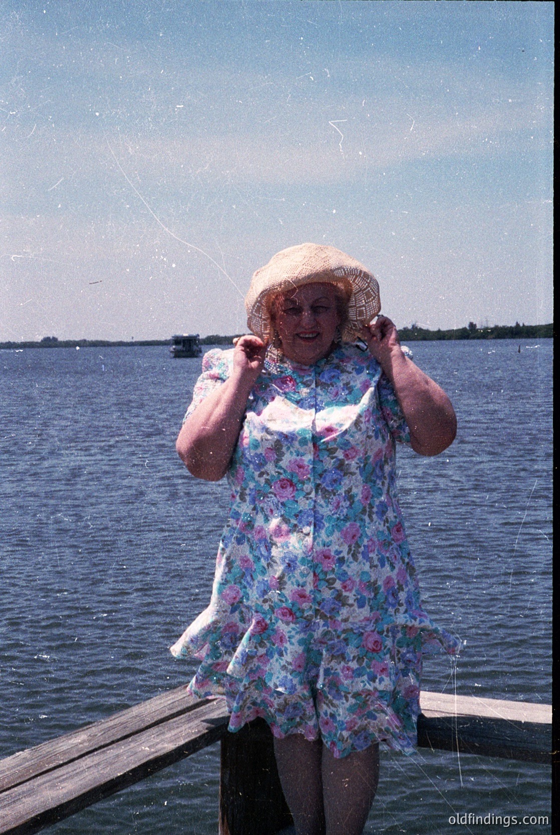 Vintage seaside portrait of a woman in a floral dress and straw hat, holding a fishing net near calm waters. Mid-20th century beachwear and fishing gear suggest recreational activity.