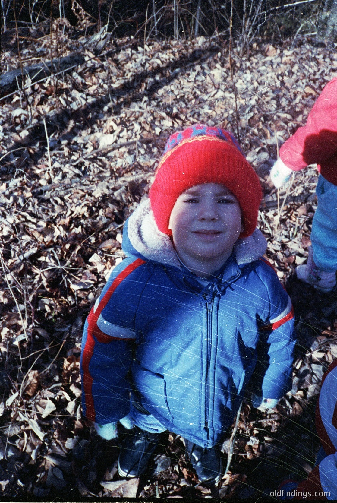 Child in winter attire—red knit hat with white trim, blue puffer jacket with red/orange stripes—playing in a wooded area covered in fallen leaves. Snow dusts their cheeks and hat. Likely late 20th century (1970s–1990s) based on clothing style. Evokes nostalgia for childhood outdoor play.
