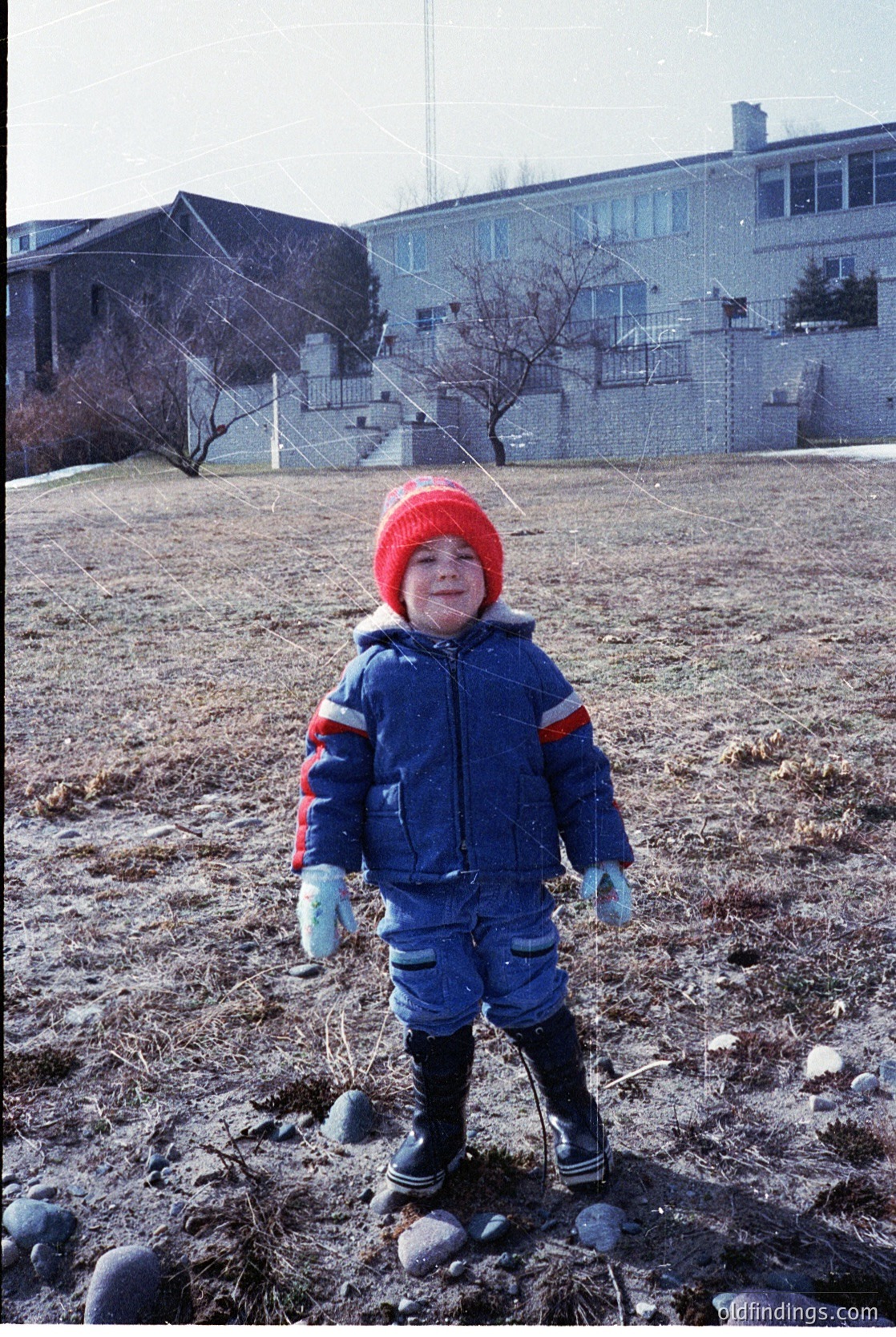 Child in winter gear—red knit hat, blue puffer jacket with red/white stripes, and black boots—standing on a grassy/sandy area with scattered rocks. Multi-story institutional building with balconies and bare trees in background. Likely 1970s–1990s outdoor schoolyard or playground setting.