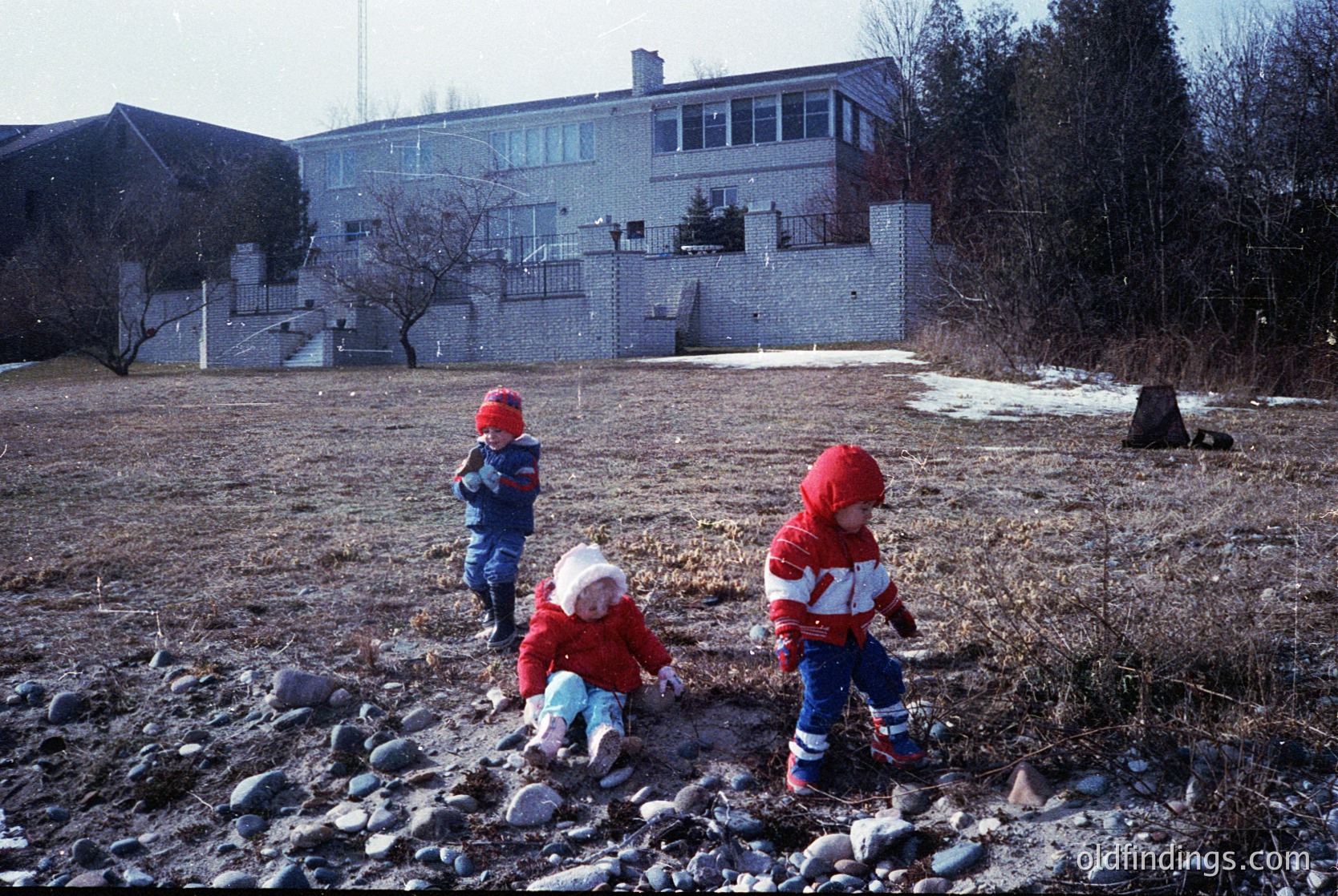 Three children in winter attire explore rocky terrain near a mid-20th-century institutional building. Snow patches and bare trees suggest late fall/winter. Clothing hints at 1960s–1970s European style.