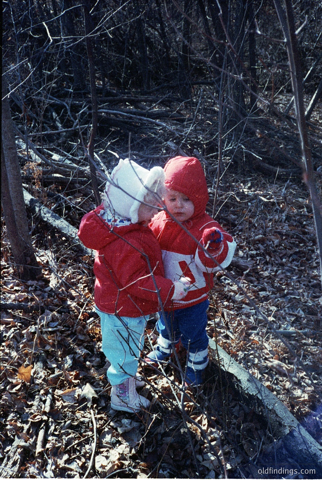 Two young children in matching red winter gear explore a wooded forest floor, likely late 20th century. The child on the left wears a white hat and light blue pants, while the other sports a red knit hat and striped boots. Branches and fallen leaves dominate the natural setting.