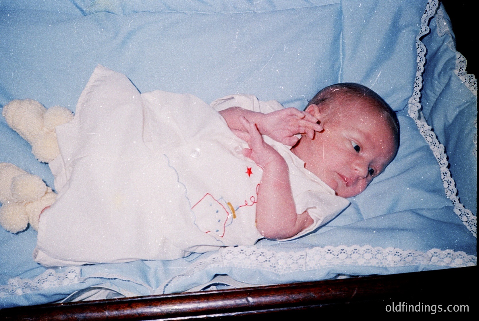 Vintage newborn portrait in a vintage-style wooden crib with lace trim, dressed in a white onesie with a cartoon character appliqué. Soft focus and warm lighting evoke a nostalgic 1990s-2000s family album feel.