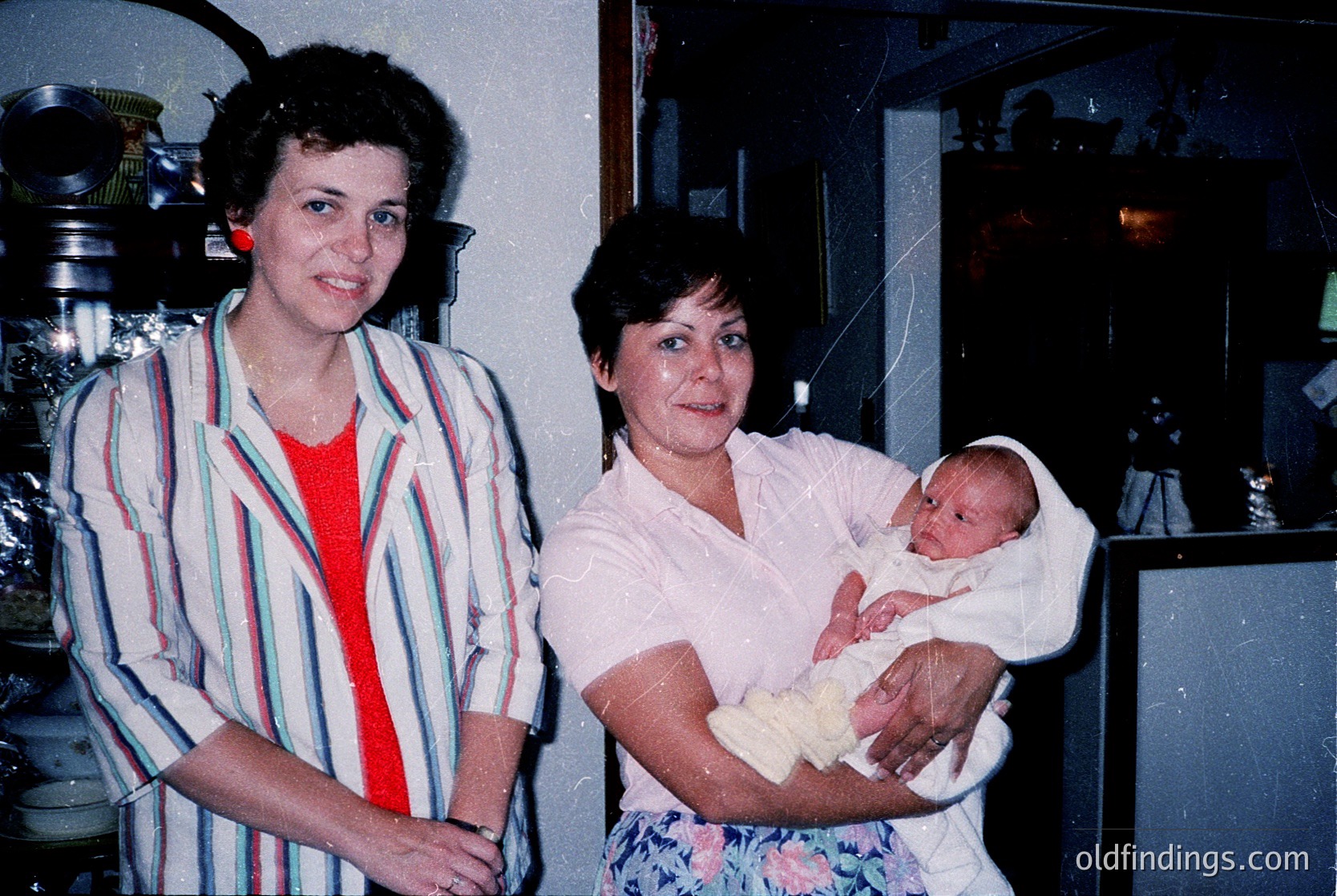 Vintage indoor portrait featuring two women and a newborn in a domestic setting, likely mid-1970s–1980s. The woman on the left wears a striped blouse with a red undershirt; the right holds a swaddled infant. Background includes vintage kitchen appliances and floral-patterned wallpaper.