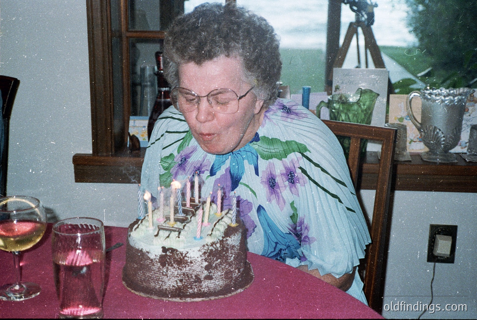 A woman in a floral-patterned blouse blows out lit candles on a round chocolate birthday cake with "Happy Birthday" written in frosting. Glasses of wine and a pitcher sit on a red tablecloth beside her. Indoor setting with wooden furniture and a view of greenery outside.
