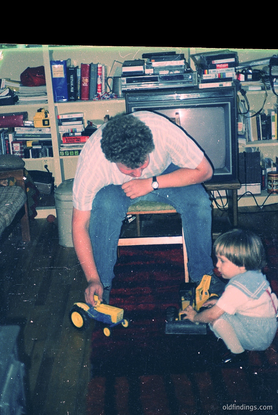 Vintage indoor scene featuring a man (mid-40s) kneeling beside a child (toddler) playing with a yellow toy truck on a wooden floor. Surrounding them: retro CRT TV, shelves stocked with books, cassettes, and vinyl records. Mid-20th century domestic setting, likely 1970s–1980s.