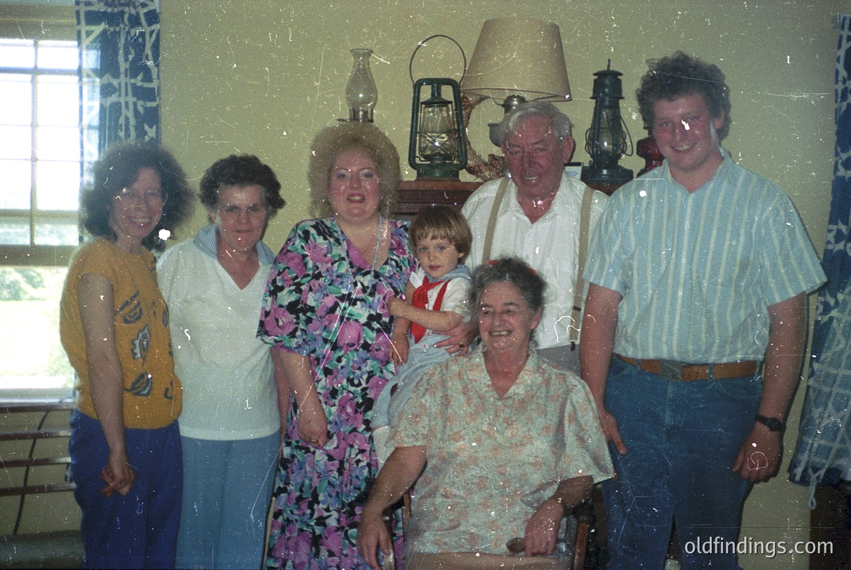 Family portrait from the 1970s–1980s, featuring seven individuals in a dimly lit indoor setting. Floral patterns dominate attire, with a vintage lantern and patterned wallpaper adding context. Smiling expressions and casual poses suggest a celebratory or informal gathering.