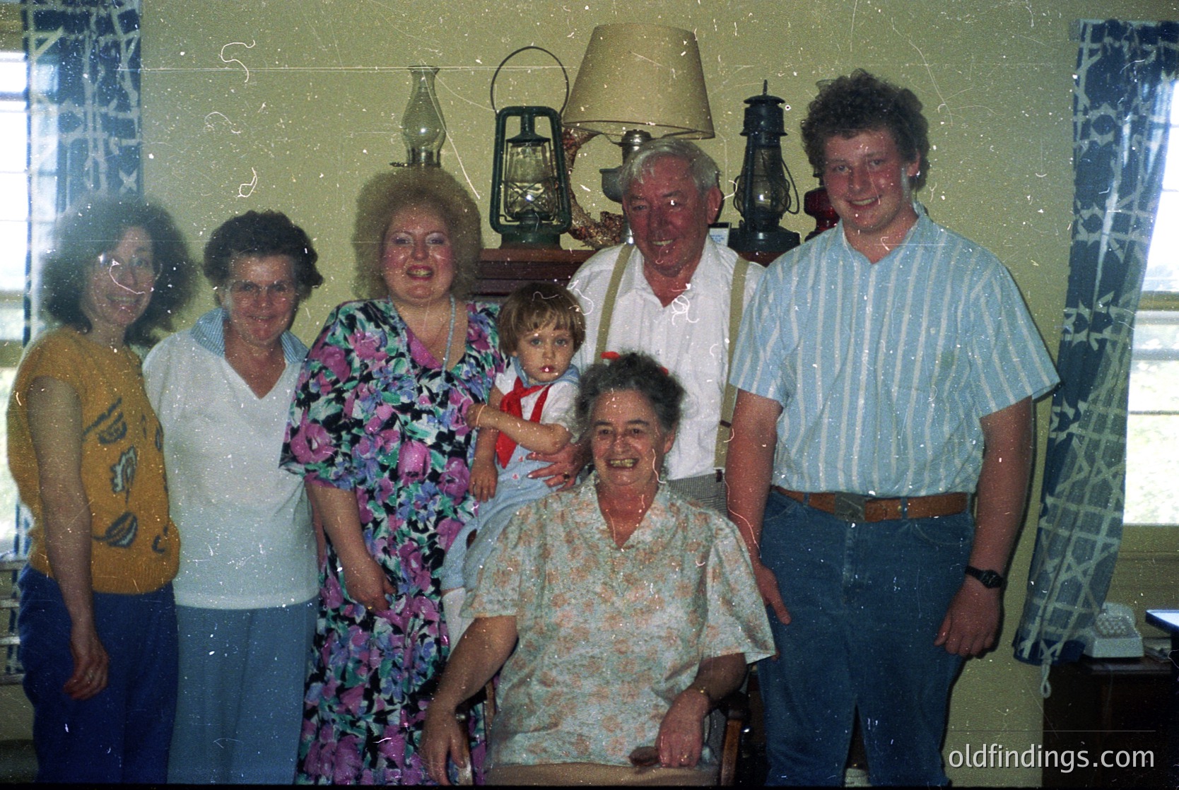 Family portrait from the 1980s–1990s featuring seven individuals in a dimly lit indoor setting. Central figure kneels holding a toddler; others stand behind in vintage attire—floral prints, striped shirts, and patterned jackets. Decor includes wall-mounted lanterns and patterned wallpaper. Warm, nostalgic atmosphere.