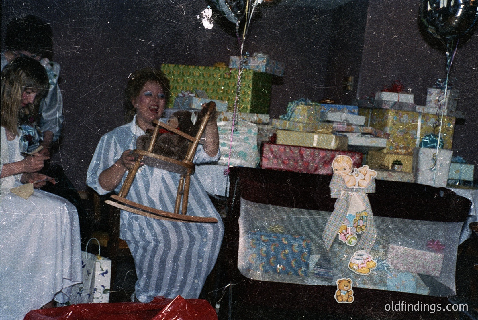 Vintage holiday scene featuring a woman in a striped apron holding a wooden rocking horse. Surrounding her are wrapped gifts, a ceramic figurine, and a tablecloth with holiday-themed designs. Warm, nostalgic lighting suggests a festive indoor gathering, likely a 1970s-1980s Christmas or holiday party.