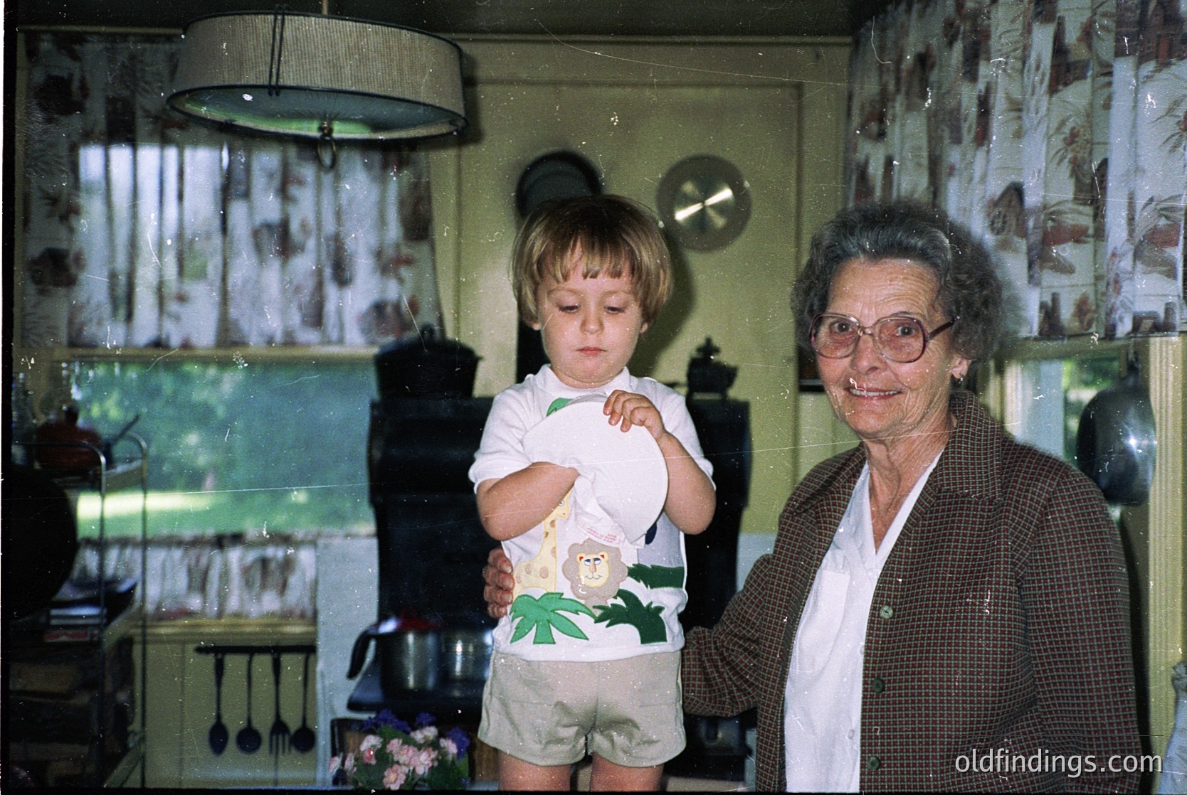 Vintage kitchen scene featuring a grandmother and toddler in mid-20th century home. Woman in patterned blazer poses beside child holding a stuffed animal. Green walls, wallpapered shelves, and retro light fixture visible.