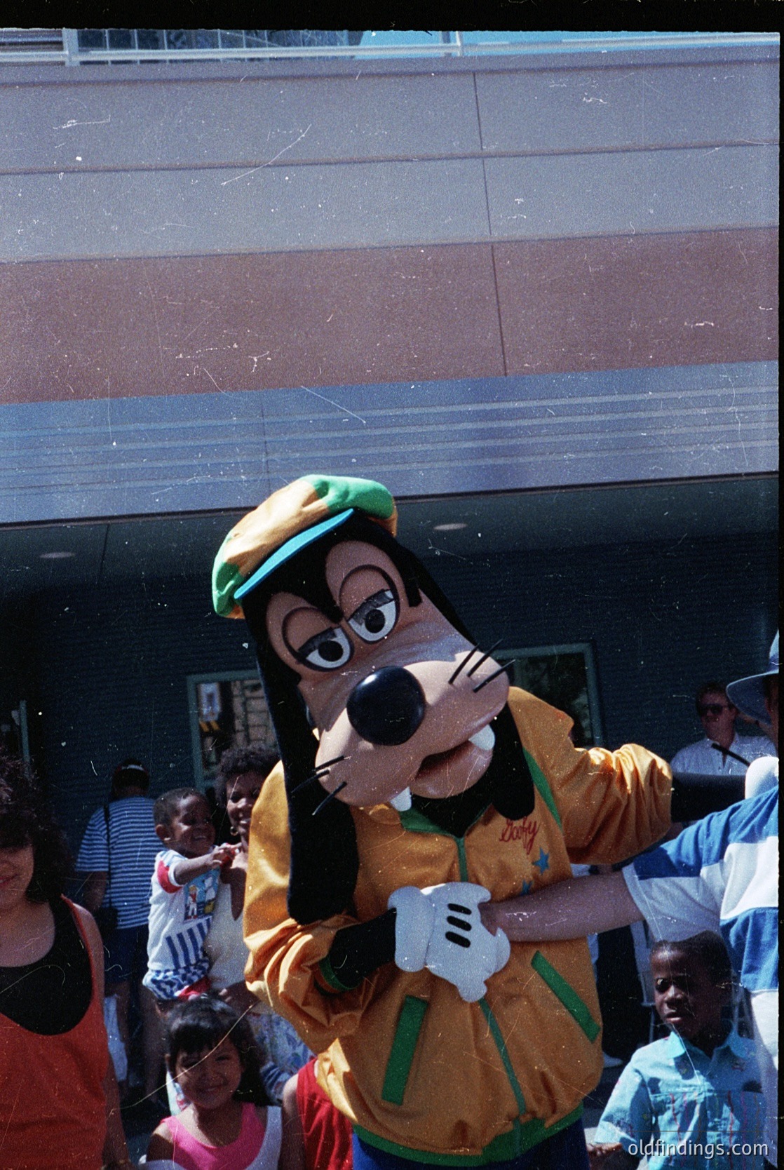 Goofy mascot in vintage Disney costume (yellow shirt, green hat) engaging with crowd at outdoor event. Reflective glass facade and modern architecture in background. Likely 1990s–2000s promotional event.