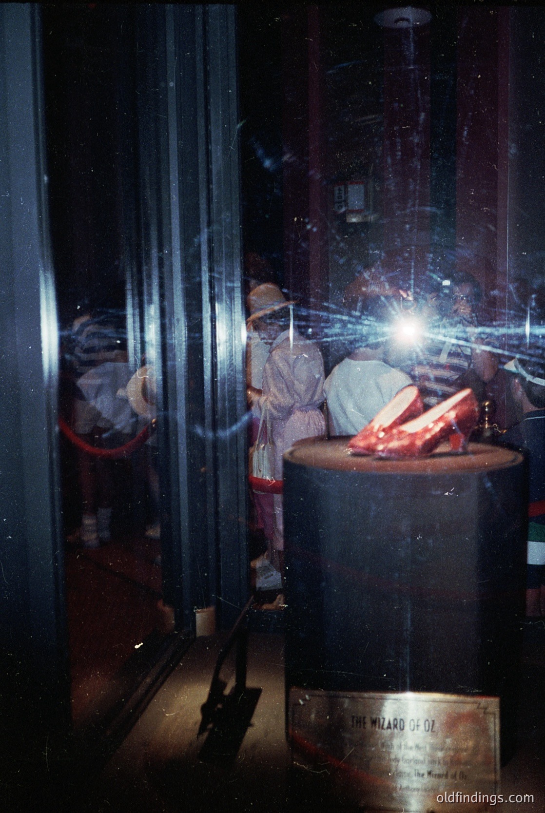 Vintage theatrical display of Dorothy’s iconic ruby slippers from *The Wizard of Oz* (1939), showcasing intricate craftsmanship and reflective glass case. Costumed figures in period attire pose beside the exhibit, emphasizing mid-20th-century stage design.
