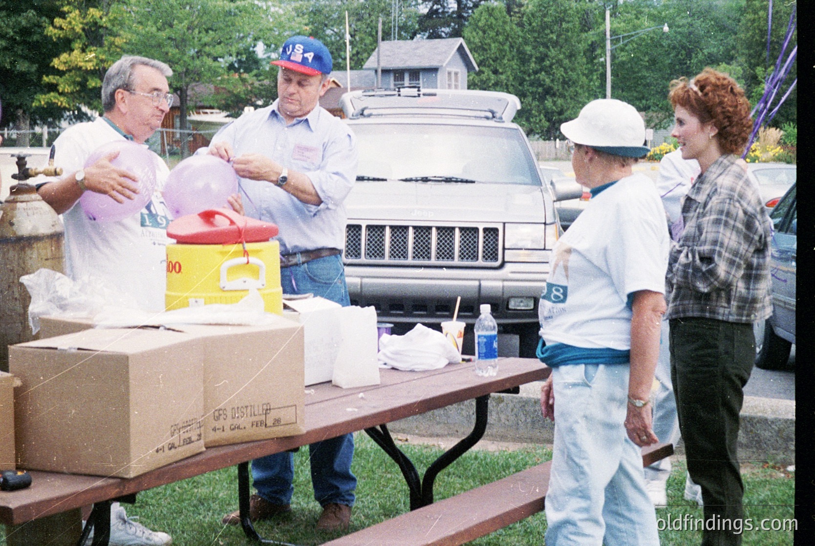 Community event setup in suburban setting, likely 1990s. Volunteers organize supplies: red cooler, yellow container, and cardboard boxes labeled "CPS" on a picnic table. Two men in baseball caps and white shirts, one woman in a white hat and plaid shirt, another in light blue jeans and cap. White pickup truck and residential homes in background.