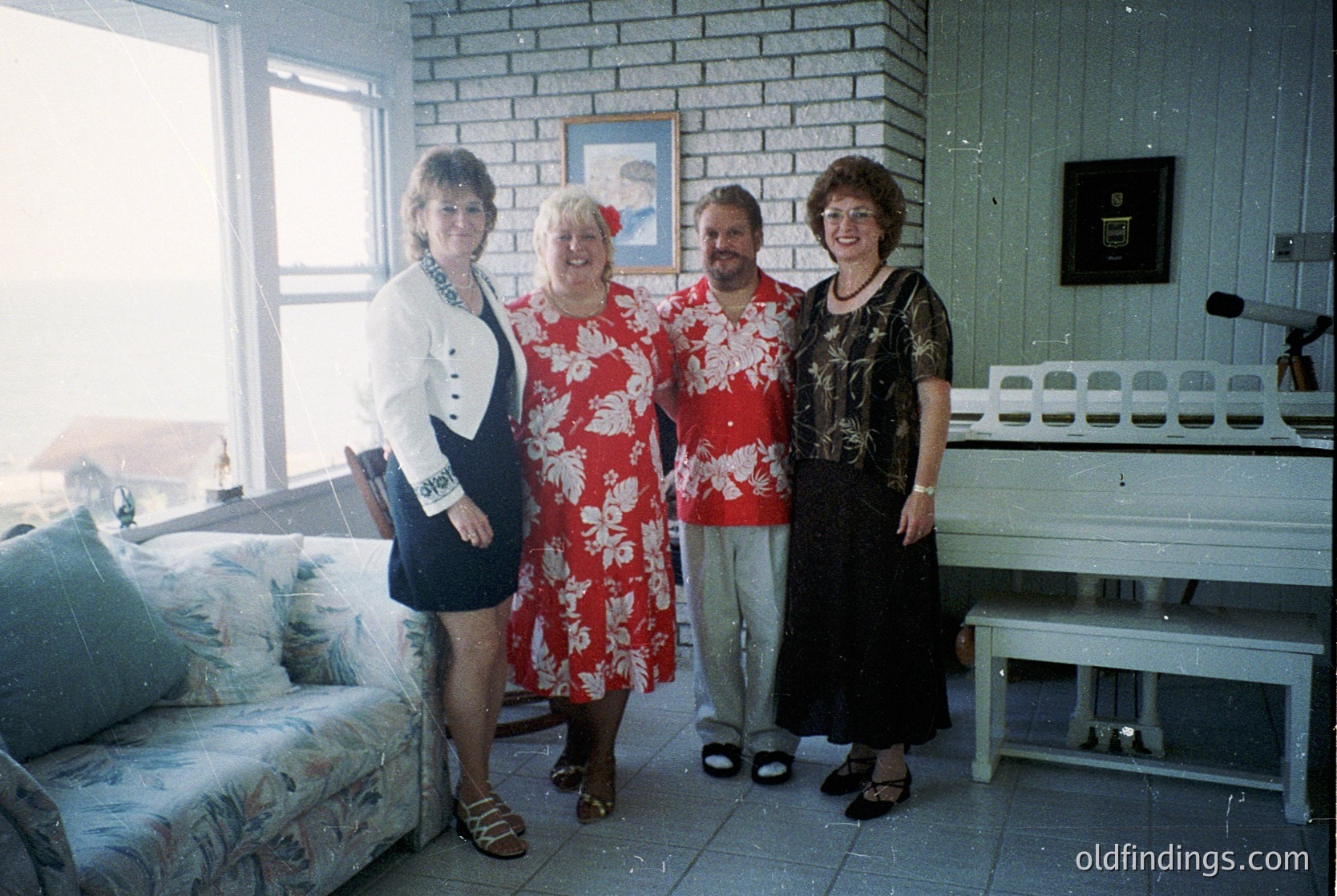 Four individuals pose indoors in a mid-century interior, likely 1970s–1980s. The woman on the left wears a structured blazer with a navy skirt; the central figure in a floral print dress; the man in a red floral shirt; the woman on the right in a black lace dress. Brick walls, a sofa with floral upholstery, and a vintage radiator define the space.