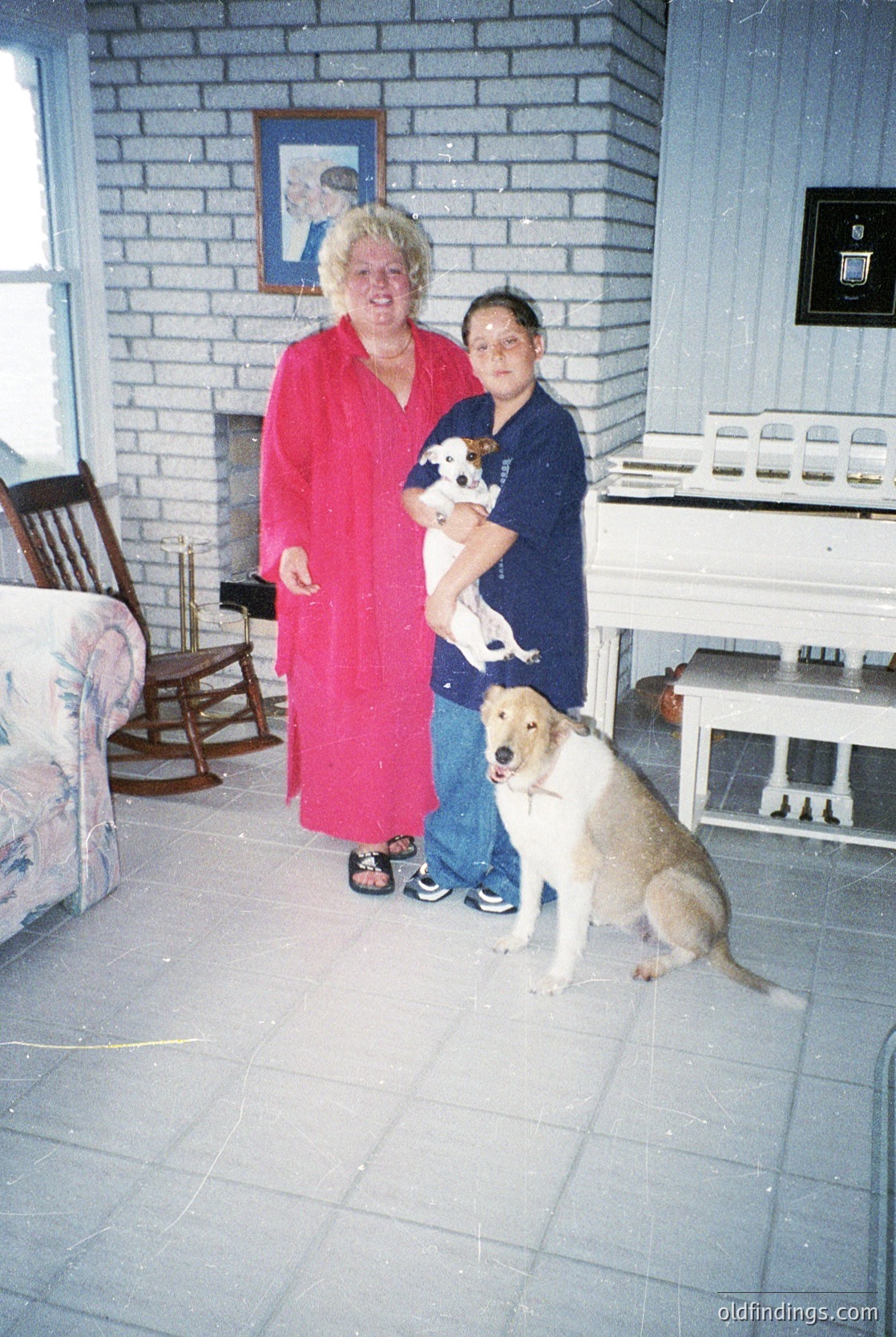 Two women pose indoors with dogs in a brick-walled room, likely mid-20th century. The woman in red wears a long robe; the woman in blue holds a small dog, while a larger dog sits beside her. Wooden furniture and floral-patterned upholstery suggest a domestic setting.