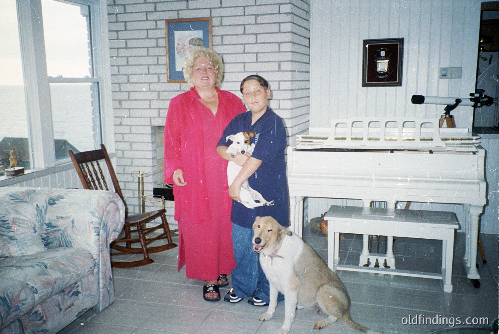 Vintage indoor portrait featuring two women and two dogs in a brick-walled room with 1990s-era decor. One woman wears a red robe, the other a blue apron; a small dog sits on her lap, while a larger dog stands beside them. Wooden cabinetry and a vintage stove suggest a domestic kitchen/living space. Warm lighting enhances nostalgic atmosphere.